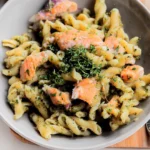 Penne pasta cooked in creamy pesto sauce, shown before and after adding grilled salmon chunks in a skillet