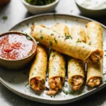 A plate piled high with golden brown, authentic Crispy Chicken Taquitos served with guacamole and salsa.